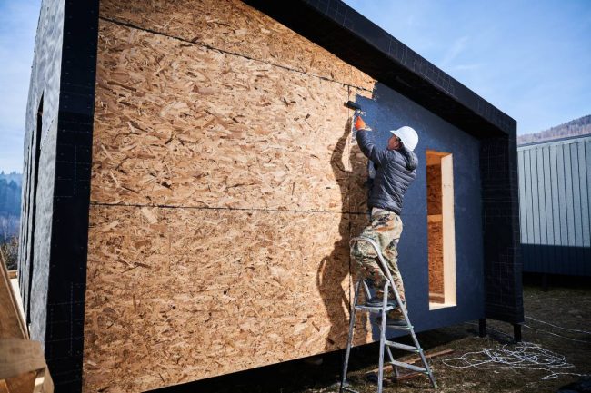 Male painter using paint roller, doing exterior paint work in a black color, standing on a ladder. Man worker building wooden frame house. Carpentry and construction concept.