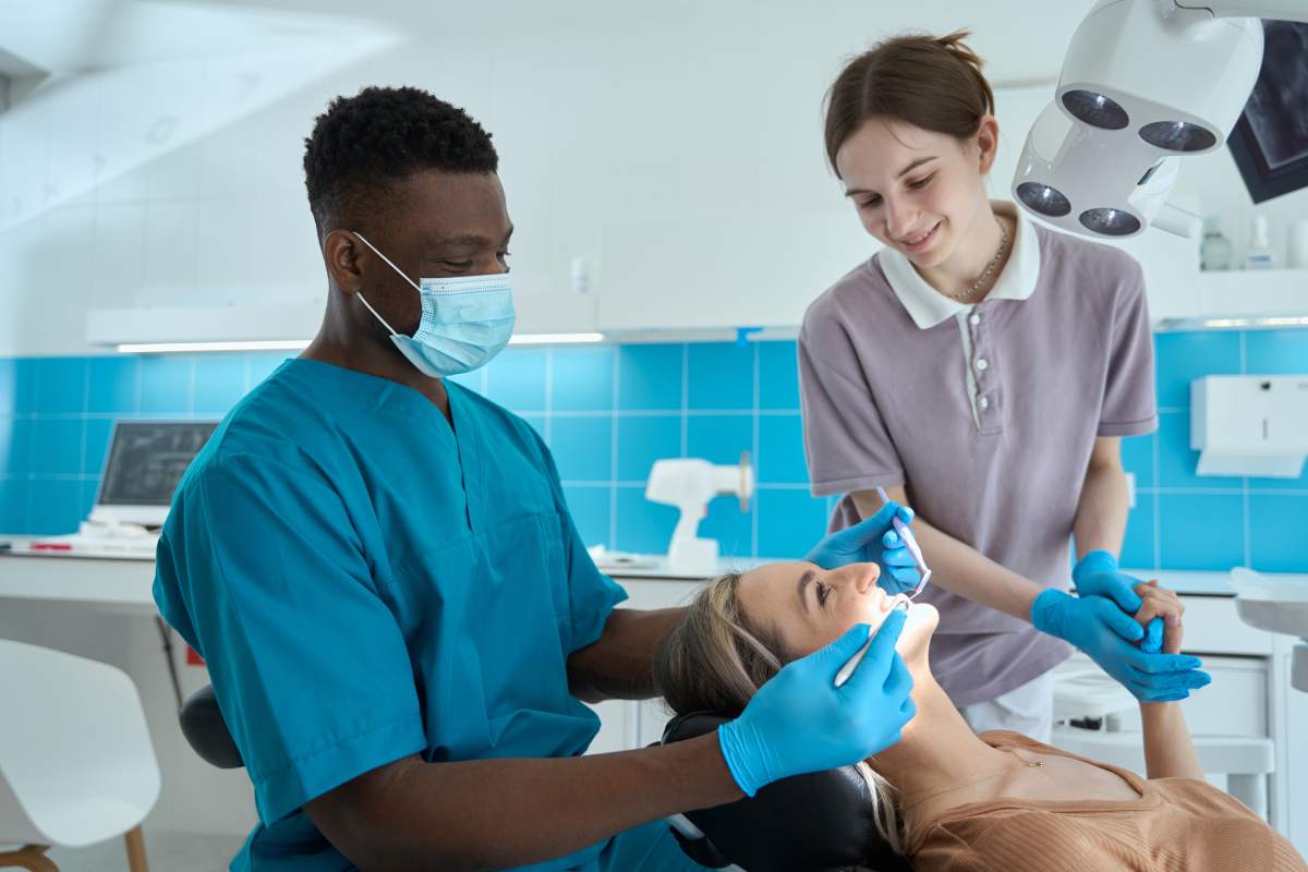 Male in protective mask and blue suit sitting on chair near female and holding dental mirror